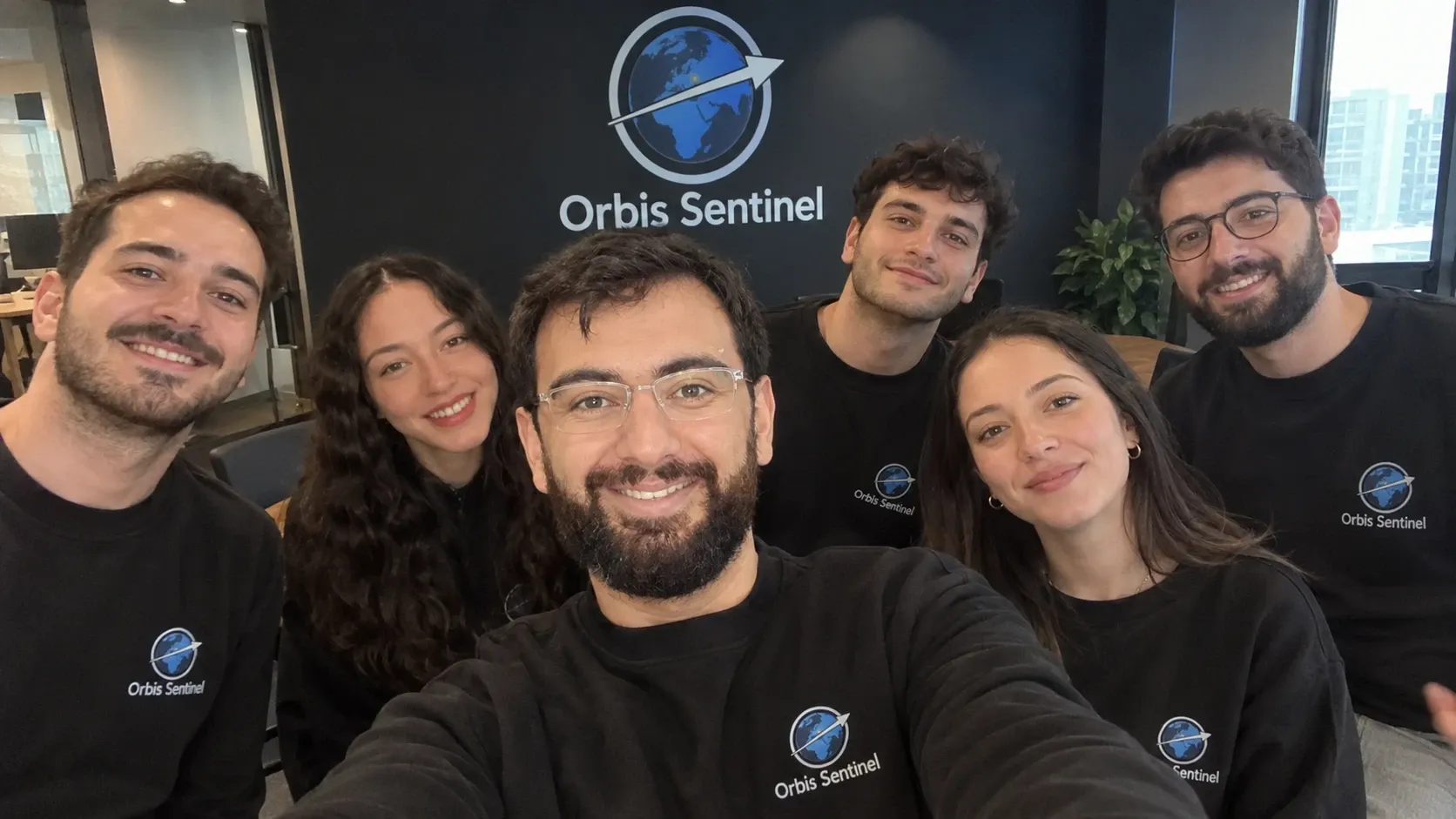 The Orbis Sentinel team, six people in black Orbis Sentinel sweatshirts, smiling in a selfie in front of the Orbis Sentinel logo in our office. Mehmet Sadık Sunay, our founder, is in front, holding the camera.
